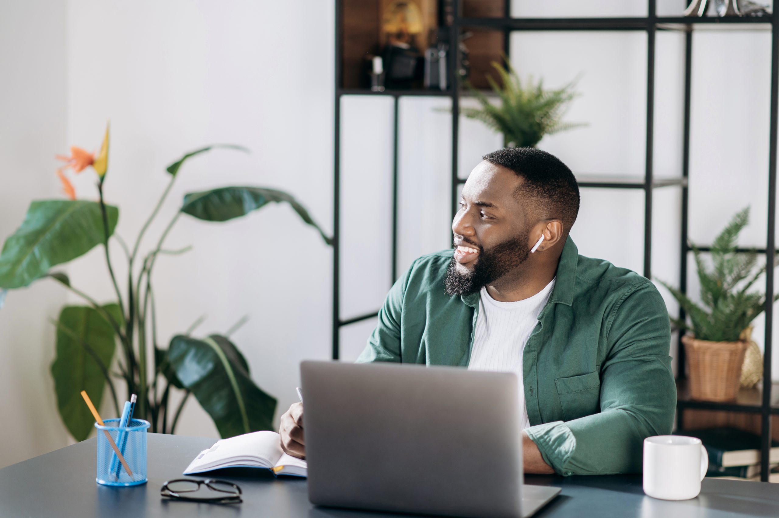 Confident african american young man sitting at work desk, using laptop for work, looking away and smiling, thinking about weekend or successful deal